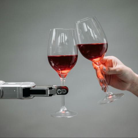 A human hand and a robotic arm clink two wine glasses filled with red wine.