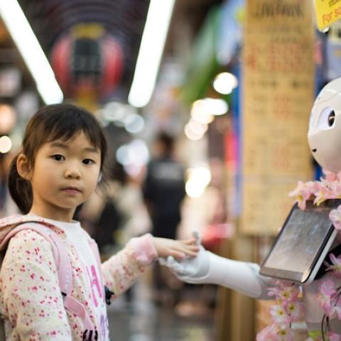 A young girl with a backpack touches the hand of a white, humanoid robot  in a busy market.
