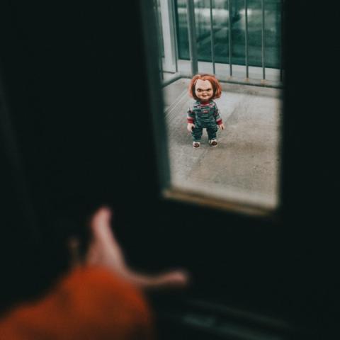 A small Chucky doll stands in a doorway, viewed through a dark, blurry frame.